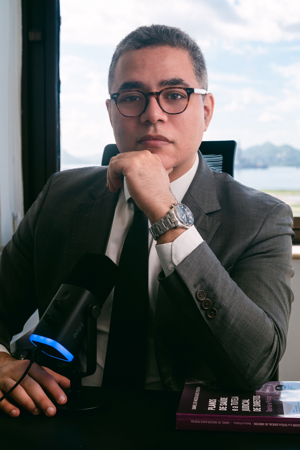 Dr. Thiago Gil, advogado previdenciário, em escritório com microfone e vista para o mar, comunicando autoridade e clareza sobre benefícios do INSS.